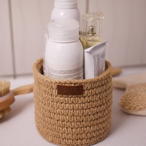 May include: A small, round, woven basket made of natural-coloured jute, holding several white bottles and tubes. A small brown leather tag is attached to the basket. The basket is designed to hold toiletries or other small items.