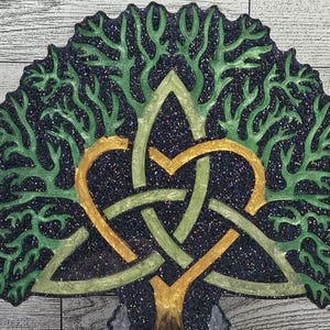 May include: A decorative tree of life design with a Celtic knot heart. The tree features green leaves and a gold and green knot against a dark, glittery background. The artwork is mounted on a wooden surface.