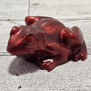 Könnte beinhalten: Dekorative Froschfigur in tiefroter Farbe mit metallischem Glanz. Die Figur sitzt auf einer hellen Holzmaserung. Ein dekoratives Objekt.
