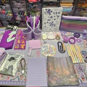 May include: A collection of purple stationery and accessories, including pens, notebooks, stickers, and a pencil case, arranged on a floral patterned desk mat. Other items include a headband, socks, and decorative paper clips.