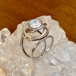 May include: A silver-toned ring featuring a multi-layered, overlapping circular band design. The top of the ring is accented with a single, round, clear faceted stone set within the center of the uppermost circular metal loop.