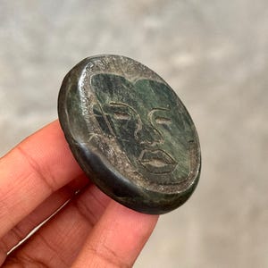 Puede incluir: Una talla de piedra ovalada de color verde oscuro que representa un rostro estilizado con los ojos cerrados y una boca definida. La talla se sostiene en una mano, destacando los intrincados detalles. La piedra tiene una superficie pulida y lisa.