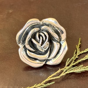 May include: A detailed, silver rose sculpture with intricate petal detailing. The rose is centered on a brown surface, with a sprig of green foliage in the lower right corner. The sculpture is a decorative item.