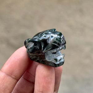 May include: A small, dark green stone carving shaped like the head of a big cat, such as a panther or jaguar. The sculpture features detailed facial carvings, including visible teeth, a snout, and ears, with a polished, glossy surface finish.