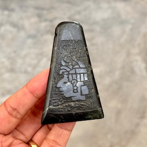 Puede incluir: Un colgante trapezoidal verde oscuro con un diseño de glifo maya tallado. El colgante se sostiene en una mano, mostrando los intrincados detalles de la antigua obra de arte. El glifo presenta un perfil de rostro con un elaborado tocado.