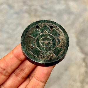 May include: A circular dark green stone pendant featuring a carved geometric pattern with a central horizontal line and two small holes at the top for suspension. The surface displays natural mineral veining and a smooth, polished finish.