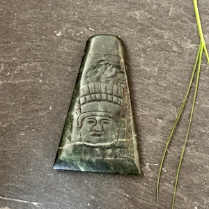 May include: A dark green, trapezoid-shaped stone carving featuring a detailed face with a headdress. The carving is set against a dark, textured background, with a few blades of green grass.