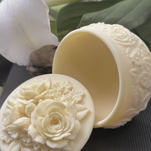 May include: Cream-coloured, oval trinket box with a floral design. The lid features a detailed relief of roses and leaves. The box's rim is adorned with a similar floral pattern. The box is open, revealing a smooth interior. A white flower is in the background.