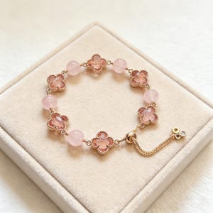 May include: A delicate rose gold bracelet featuring alternating pink quartz beads and clover-shaped charms. The bracelet has a sliding clasp for adjustable sizing and is displayed on a neutral-toned surface. The clover charms have a faceted appearance.