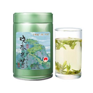 May include: A cylindrical light green metal tea canister with a label featuring a mountain landscape illustration and Chinese text, alongside a clear glass filled with light yellow tea liquid and submerged green tea leaves, net weight 50g.