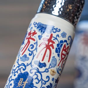 以下が含まれることがあります： 白地に青い花柄と赤い漢字が描かれたウォーターボトル。ボトルは銀色のテクスチャード加工の蓋と金のアクセントが付いています。デザインは青い花とスクロール模様が特徴です。