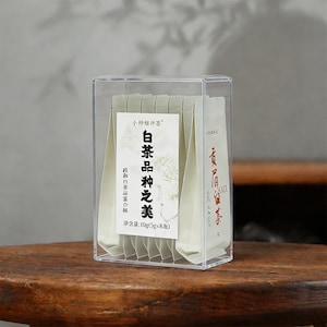 May include: A clear acrylic box containing individually wrapped tea bags. The tea bags are white with Chinese text and graphics. The box sits on a wooden surface. The text on the label reads "White Tea Varieties of Beauty".