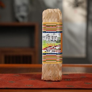 May include: A rectangular package wrapped in woven bamboo. The package has a colorful label with Chinese characters and a landscape illustration. The label includes the year 2013. The package is sitting on a red surface.