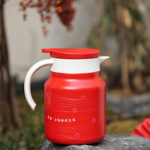 May include: A red insulated thermos with a white handle and accents. The thermos has a red lid and a white band around the middle. The thermos has a small floral design and Chinese characters. The thermos is sitting on a gray rock.