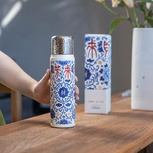 以下が含まれることがあります： 銀色の蓋が付いた、白と青の模様のサーモス。サーモスには赤と青の漢字が書かれています。同様のデザインの箱がサーモスの隣にあります。サーモスは人が持っています。