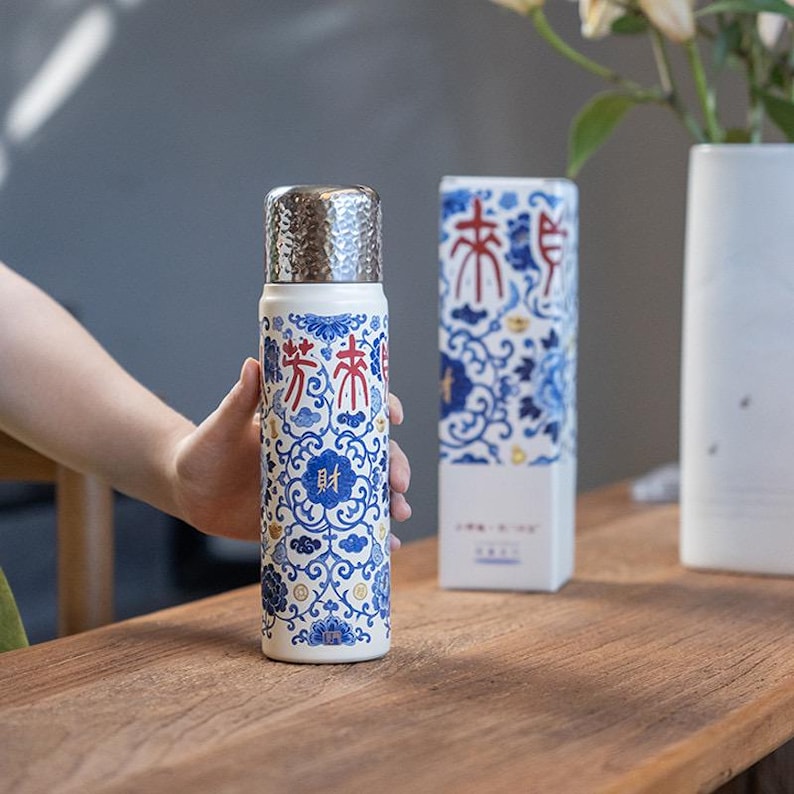 以下が含まれることがあります： 銀色の蓋が付いた、白と青の模様のサーモス。サーモスには赤と青の漢字が書かれています。同様のデザインの箱がサーモスの隣にあります。サーモスは人が持っています。
