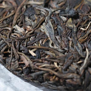 May include: Close-up of a compressed tea cake, showcasing a dense arrangement of dark brown and grey tea leaves. The texture is rough and fibrous, with visible stems and varying shades of brown. The tea cake appears to be aged.