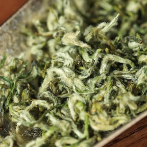 May include: Close-up of loose leaf tea with a mix of green and white hues. The tea leaves have a delicate, fuzzy texture, suggesting a high-quality product. The image highlights the natural colors and textures of the tea.