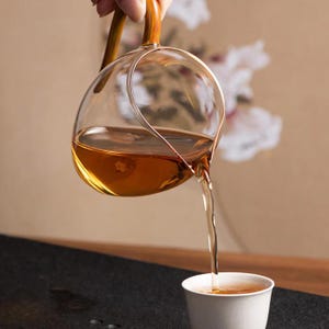 May include: A clear glass tea pitcher with a bamboo handle is pouring amber tea into a white cup. The pitcher has a rounded shape and a long spout. The background is a soft, neutral color with a floral design.