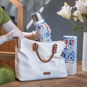 以下が含まれることがあります： 青い花柄と銀色の蓋が付いた白い断熱ウォーターボトルが、茶色の革のハンドルが付いた白いトートバッグに入れられています。同じデザインのマッチングボックスが見えます。
