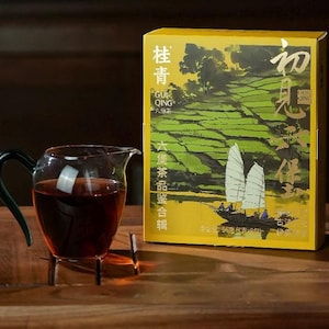 May include: A glass teapot with a dark handle and a dark liquid inside, resting on a metal stand. A yellow box with Chinese characters and a landscape illustration is behind the teapot. The box has the text "GUR QING" and "56 grams".