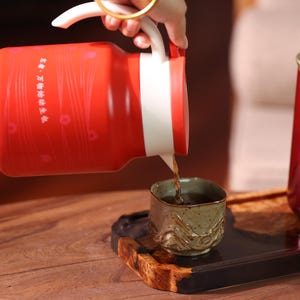May include: A red and white tea pot is pouring tea into a small, decorative cup. The teapot has a white handle and a red body with a floral design. The cup is sitting on a wooden tray.