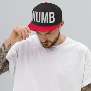 May include: A black and red baseball cap with the word "NUMB" in large white letters. The cap has a flat brim and a structured fit. The person in the image is wearing a white t-shirt.