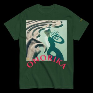 Peut inclure: T-shirt vert foncé avec un motif surréaliste dans un cadre carré. Le motif présente une figure et des formes abstraites. Le mot "ONORIKA" est imprimé en rose. Un petit motif doré est sur la manche.