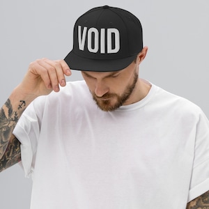 May include: Black baseball cap with the word "VOID" in bold white letters across the front. The cap has a flat brim and is worn by a person wearing a white t-shirt.