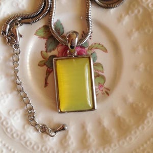 Peut inclure: Un collier avec un pendentif rectangulaire doté d'une pierre cabochon jaune clair lisse sertie dans un cadre en métal argenté poli. Le pendentif est suspendu à une chaîne serpent argentée souple avec un fermoir mousqueton et une chaînette.