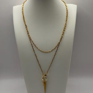 May include: A gold-colored necklace displayed on a white mannequin. The necklace features two chains, one shorter and one longer, with a pendant. The pendant is a gold-colored, textured spike with a star charm. The necklace is designed for a layered look.