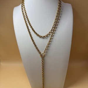 May include: A gold-toned lariat necklace featuring a long, continuous rolo chain design. The chain loops through a circular ring connector, creating a double-layered upper section and a single dangling drop that extends downwards from the centre.