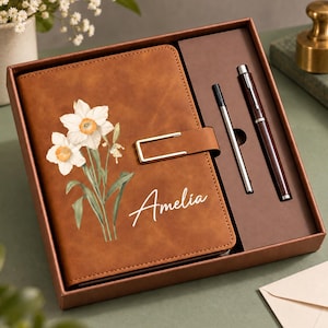 May include: A brown leather-look notebook featuring a white daffodil illustration and the name Amelia in white script, secured with a gold-tone metal clasp. The set includes a matching brown pen and a silver ink refill inside a brown gift box.
