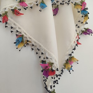 Pode incluir: Um lenço de tecido na cor creme com uma borda de crochê branca adornada com pequenas contas pretas e delicadas flores de tecido multicoloridas em tons de azul, amarelo, vermelho, roxo e rosa penduradas nas bordas.