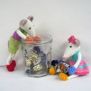 May include: Two knitted mice, one peering into a glass jar of wrapped sweets, the other sitting nearby. The mice are white with pink, green, purple, and red details. The jar is clear, and the sweets are in gold and blue wrappers.