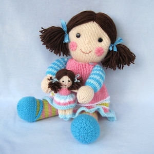 May include: Two knitted dolls, one larger than the other, are sitting together. The larger doll has brown hair, blue and white striped sleeves, and a pink dress. The smaller doll has brown hair, a pink dress with white stripes, and is being held by the larger doll.