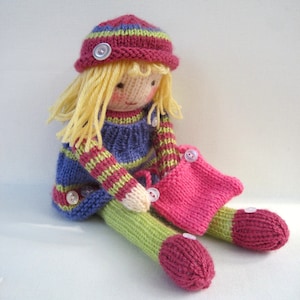 May include: A knitted doll with blonde hair, wearing a pink and green striped dress, a pink hat with green stripes, and pink shoes. The doll is holding a pink knitted bag.