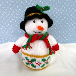 May include: A white knitted snowman wearing a black hat with a green holly sprig, a red and green scarf, and red mittens. The snowman has a carrot nose and black eyes.