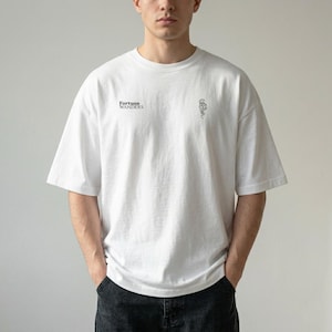 Può includere: T-shirt bianco oversize a maniche corte. La maglietta presenta la scritta "Fortune Wanders" sul petto sinistro e una piccola grafica sul lato destro. Il modello indossa jeans neri.