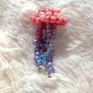 May include: A handmade beaded jellyfish in shades of pink, purple, and blue. The jellyfish has a rounded top and dangling tentacles, with a small, clear ring at the bottom. The beads are various sizes and shapes.