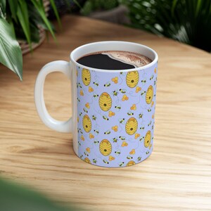 May include: White ceramic coffee mug with a handle, filled with dark liquid. The mug features a repeating pattern of yellow beehives, bees, and honeycombs on a light purple background. The mug is sitting on a wooden surface.