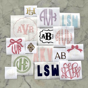 Könnte beinhalten: Eine Sammlung von Monogramm-Aufklebern in verschiedenen Farben und Stilen. Die Aufkleber zeigen Initialen in verschiedenen Schriftarten, darunter Schreibschrift und Blockbuchstaben, mit Schleifen und dekorativen Rahmen. Farben: Rosa, Gold, Grün und Marineblau.