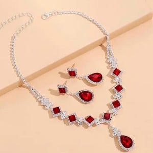 May include: A silver-toned necklace and earring set. The necklace features a series of square and teardrop-shaped red gemstones, interspersed with clear crystals. The matching earrings have a similar design, with square studs and teardrop pendants.