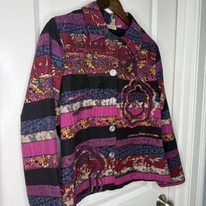 May include: A patchwork jacket with a vibrant mix of colors and patterns. The jacket features a collared neckline, button-up front, and long sleeves. The design incorporates shades of pink, purple, black, and various floral and geometric prints. Silver buttons add a touch of contrast.
