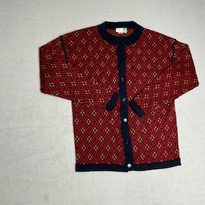 May include: A red cardigan with a geometric pattern in yellow and black. The sweater has a round, navy blue collar and navy blue trim along the button closure and cuffs. The cardigan is buttoned up.