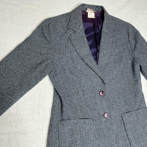 May include: A gray blazer with a subtle checkered pattern. The blazer has a purple lining, two front pockets, and two purple buttons. The blazer's sleeves are long, and the garment appears to be made of a woven fabric.
