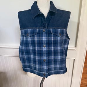 May include: A denim and plaid vest. The top portion is a dark blue denim with a collar and button closure. The lower portion is a blue plaid pattern with two pockets and button closure. The vest is displayed on a mannequin.
