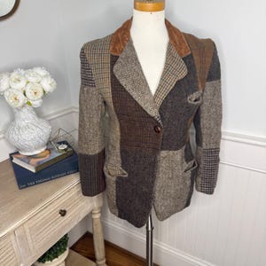 May include: A patchwork blazer featuring various brown, tan, and grey wool-like fabric panels with herringbone, houndstooth, and plaid patterns. The jacket includes a brown velvet collar, a single front button closure, and two side pockets.