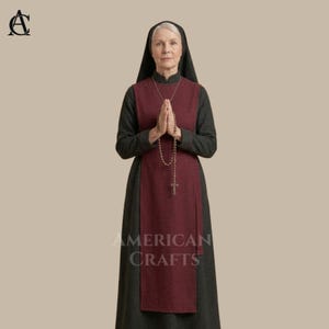 May include: A woman wearing a traditional nun's habit, featuring a black veil and a long, dark gray dress. A burgundy apron-like garment is worn over the dress. A rosary hangs around the neck. The text "AMERICAN CRAFTS" is visible.