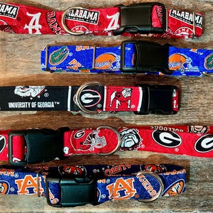 May include: Five dog collars with team logos and colors. The collars feature the University of Alabama, University of Florida, University of Georgia, and Auburn University. Each collar has a black plastic buckle and a metal D-ring.